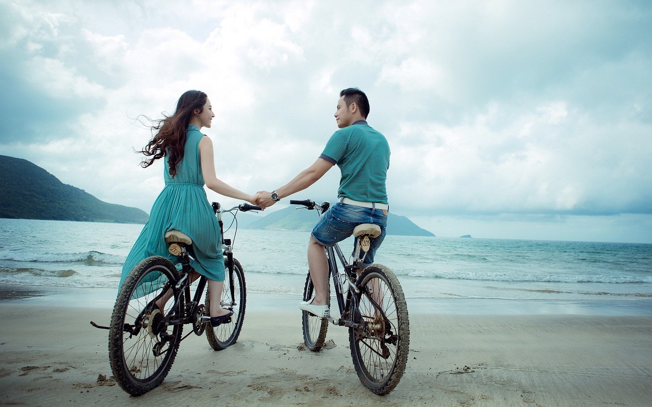 young couple on bicycle holding hands, beach side